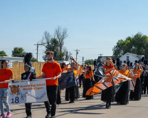 A high school marching band and color guard march down a street in a parade.