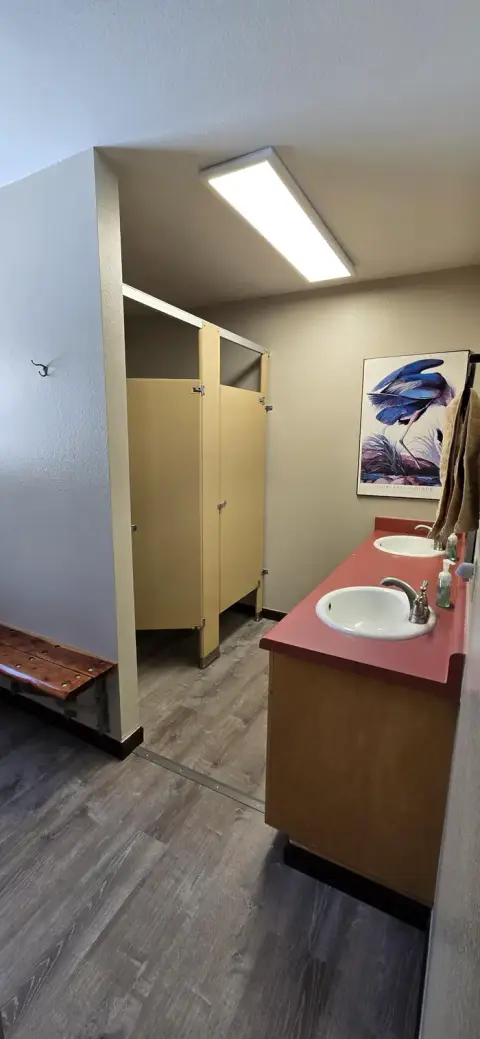 A public restroom interior features stall doors, a vanity with two sinks, and a framed bird print on the wall.