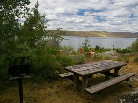 A wooden picnic table and grill sit near a lake surrounded by trees and mountains.