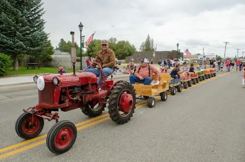 A red tractor pulls a string of small wooden wagons down a street during a parade.