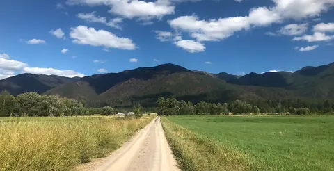 A dirt road leading toward mountains across fields under a partly cloudy sky.