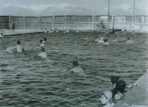 Historic black and white photograph of people swimming in an outdoor pool with mountains in the background.