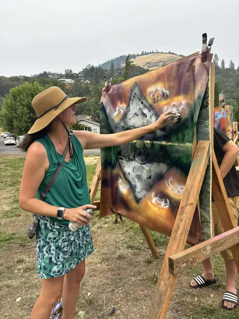 A woman creates a spray paint art piece outdoors on a wooden easel.