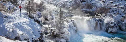 A person hiking along a snowy trail next to a frozen waterfall and river.
