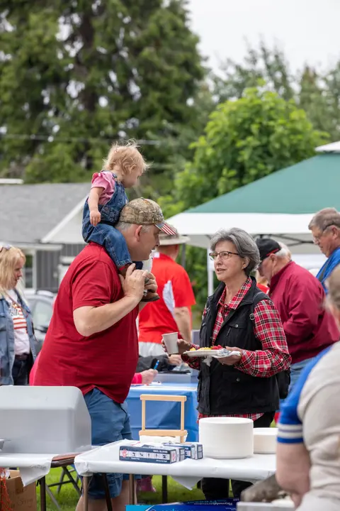 A man with a child on his shoulders talks to a woman at an outdoor gathering.