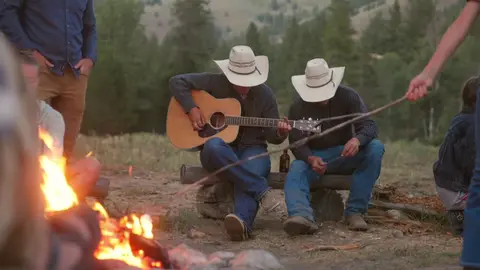 People sitting around a campfire, with two men wearing cowboy hats, one playing a guitar.