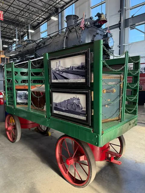A vintage baggage cart with framed historic photographs inside a museum with a steam locomotive.