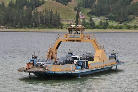 A car ferry navigates across a body of water with hills in the background.