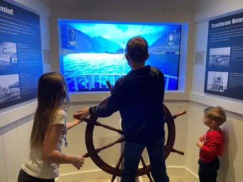 Children interact with a ship's wheel exhibit in an indoor museum space.