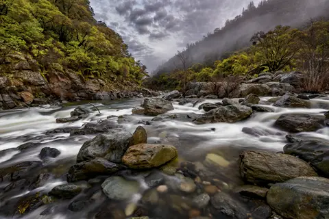 A rushing river flows through a rocky canyon with misty mountains in the background.
