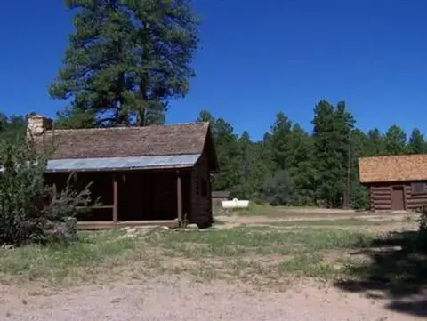 Two log cabins stand in a clearing surrounded by trees under a clear blue sky.