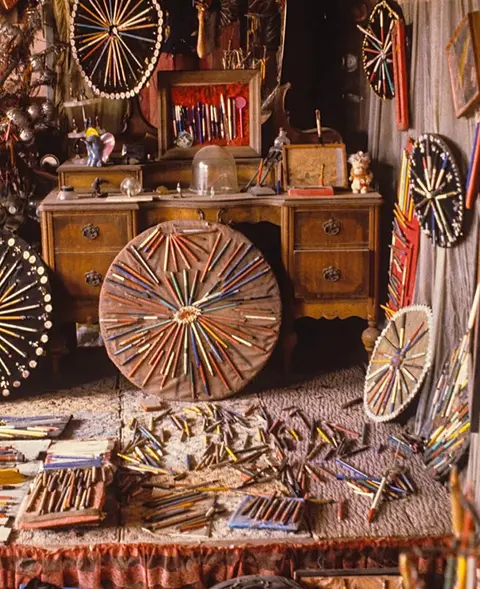 A cluttered room features a wooden desk and numerous circular arrangements of pencils.
