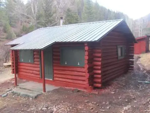 A red log cabin with a metal roof stands in a wooded mountain area.