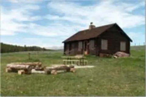 A wooden cabin stands in a grassy field with a picnic table and log seating.