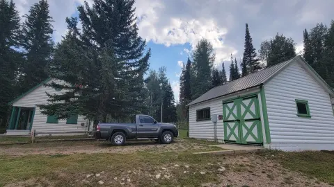 A pickup truck parked between two white wooden cabins with green trim near tall trees.