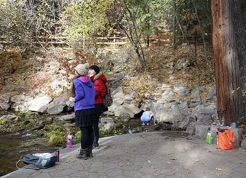 Two people stand near a rocky stream in a wooded area.
