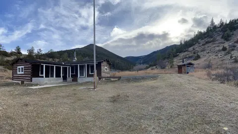A rustic wooden cabin sits in a dry grassy landscape under a cloudy sky.