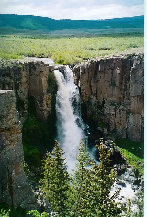 A waterfall flows through a rocky canyon with green trees and hills in the background.