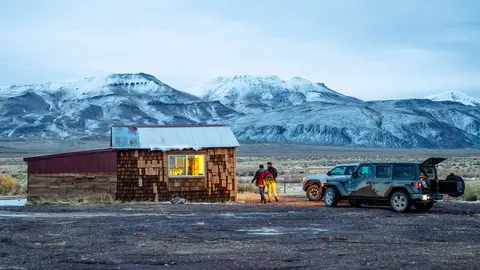 A rustic wooden cabin sits beneath snow-covered mountains with parked vehicles nearby.