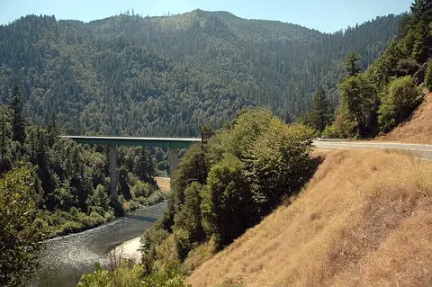 A green highway bridge spans a river through a dense forested mountainous landscape.