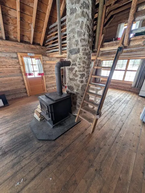 An interior view of a rustic wooden cabin showing a stone chimney, wood stove, and loft ladder.