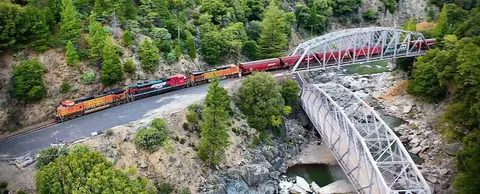 A train crosses a bridge over a river surrounded by lush green forests and rocky terrain.