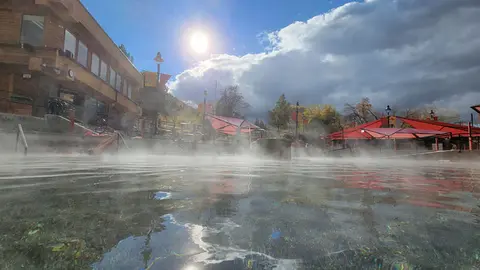 A steaming hot spring pool with a building and umbrellas in the background under a sunny sky.