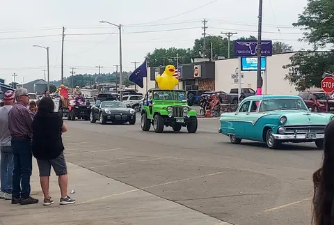 People watch a parade featuring various vehicles including a jeep with a giant yellow duck on top.