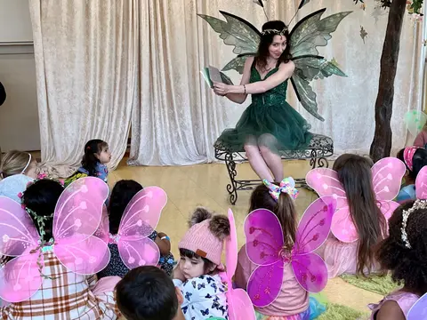 A person dressed as a fairy reads a book to a group of children wearing fairy wings.