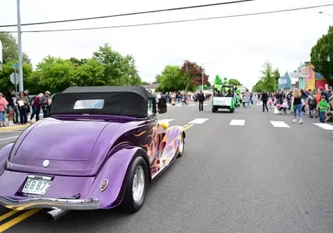 A purple vintage car with flames is parked on a street during a parade.