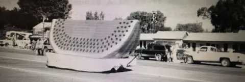 A black and white photograph showing a large watermelon slice float parked on a street.