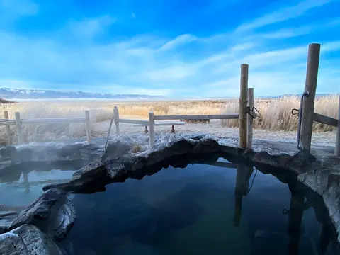 A stone hot spring pool sits outdoors overlooking a vast landscape under a blue sky.