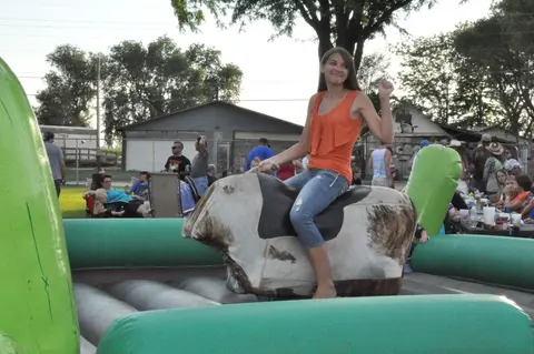A young woman rides a mechanical bull at an outdoor event.