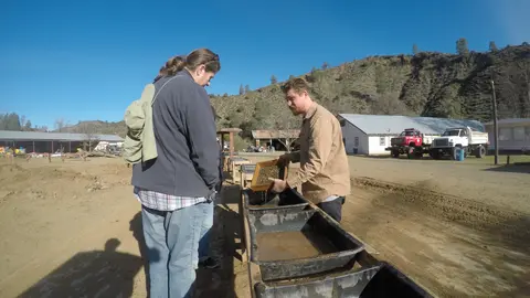 People panning for gold outdoors near troughs and buildings.