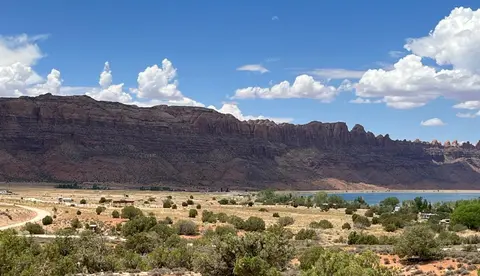 A scenic view of a desert landscape with cliffs and a lake under a cloudy blue sky.