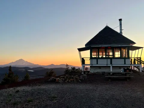 A fire lookout tower stands on a ridge at sunset with mountains in the background.