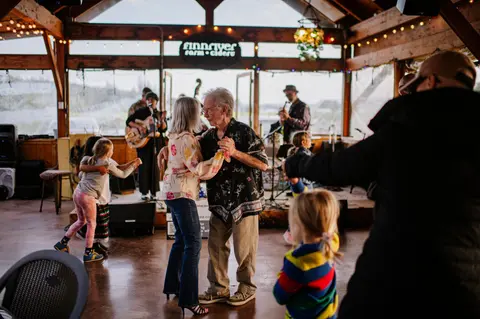 An older couple dances inside a rustic venue while children dance and a band plays in the background.