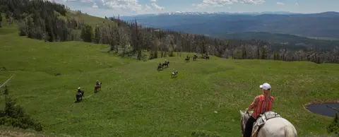 A group of people ride horses across a grassy ridge with mountains in the background.