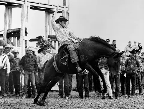 A cowboy riding a bucking horse at a rodeo while spectators watch.