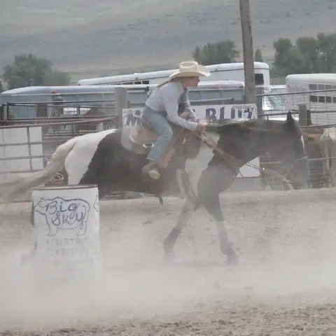 A cowgirl rides a pinto horse around a barrel in an arena.