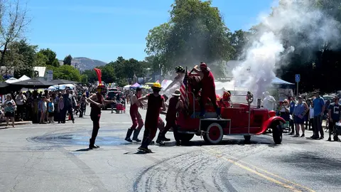 People dressed as firefighters perform near a vintage fire truck in the street.