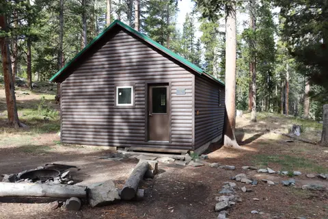 A log cabin sits in a forest clearing next to a fire pit.