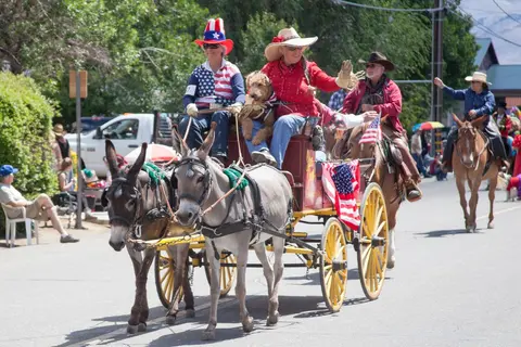 People in a parade riding on a cart pulled by donkeys and on horseback.
