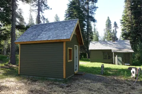Two small green wooden buildings sit in a forest clearing.