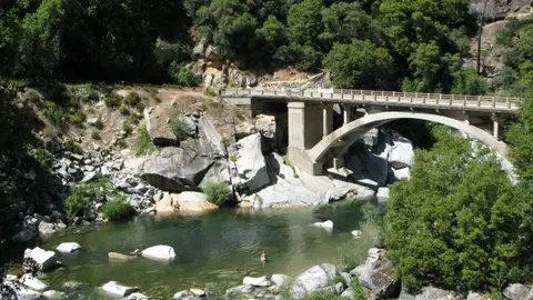A bridge spans a rocky river surrounded by trees, with people swimming in the water.