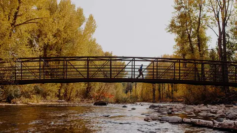 A person walks across a metal bridge over a river surrounded by autumn trees.