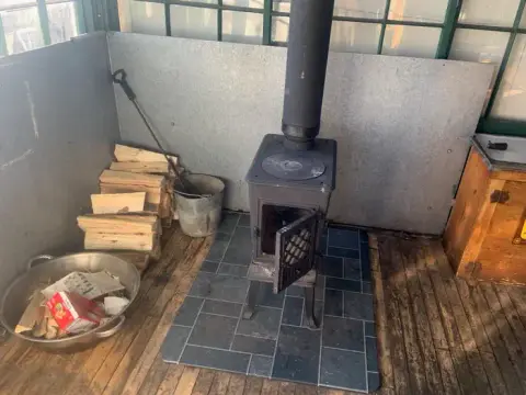 A small wood-burning stove sits on a tile hearth inside a room with wood floors and stacked firewood.