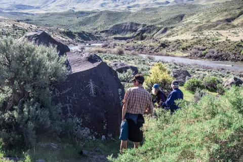 People looking at petroglyphs on a large rock by a river.