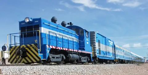 A blue locomotive and train on tracks against a blue sky.