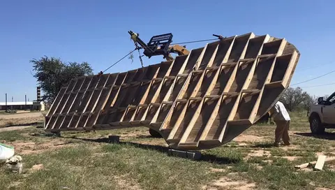 A large wooden structure is lifted by equipment outdoors.